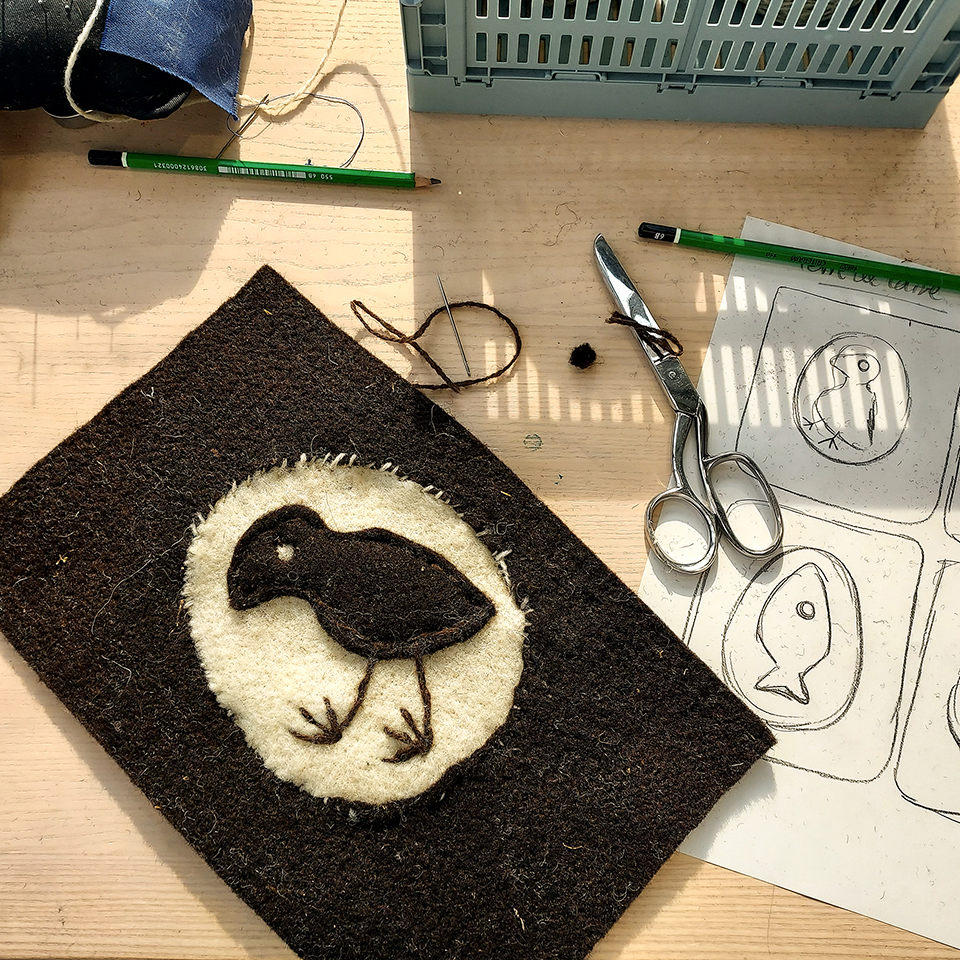A photograph of a workbench featuring several items related to felt work, including scissors, needle and thread. There are also pencils and a paper sheet with some sketches. On the left of the picture there's a felt arwork made of dark brown and off white felt featuring a childish-looking embroidery of a bird with long legs inside of an egg.